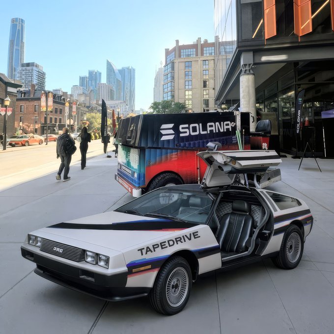 A silver DeLorean-style car with "TAPEDRIVE" written on the side and its gull-wing doors open is parked on a city sidewalk, with a "SOLANA" branded booth visible in the background near Chicago skyline buildings.