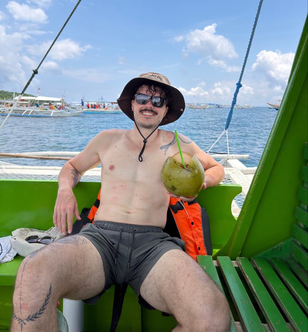 A young man wearing a wide-brimmed hat and sunglasses smiles while holding a fresh coconut aboard a green outrigger boat in the Philippines, embodying the relaxed island lifestyle that complements his bio as a German founder turned marketing lead based there.