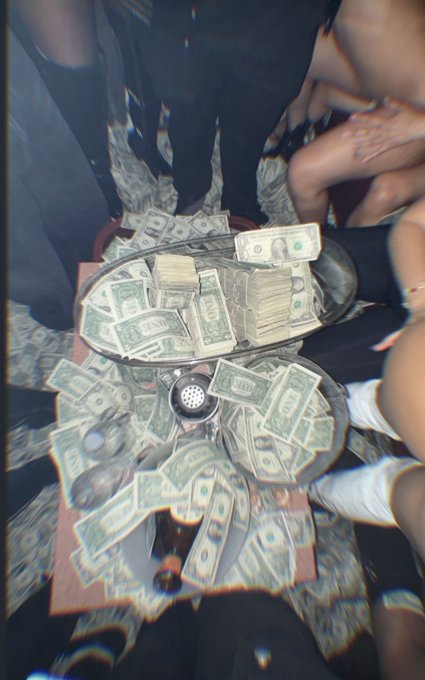 An overhead view captures a silver platter piled with stacks of US dollar bills, surrounded by scattered cash and people's legs at what appears to be a lavish party.