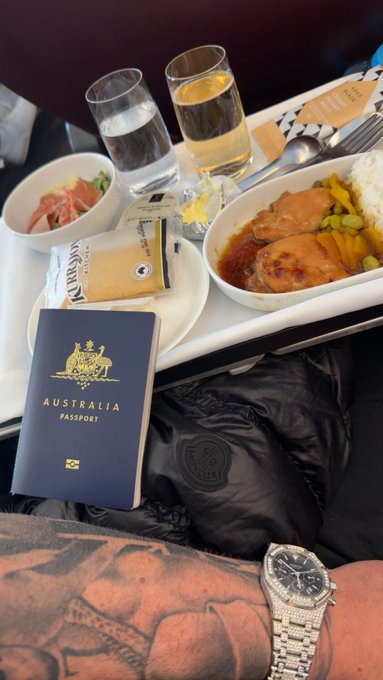 A passenger enjoys a premium in-flight meal in business class, featuring a bowl of glazed chicken with rice and vegetables, a side salad, and drinks, accompanied by a chocolate "Magnum" ice cream bar, an Australian passport, and a luxurious watch resting on a tattooed leg. The image perfectly illustrates the post's message about the perks of flying business class, where the extra space and privacy mean no awkward seating arrangements with oversized neighbors.