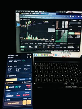 A dimly lit workspace showing a laptop displaying cryptocurrency trading charts on the TradingView platform alongside a smartphone showing a trading app interface.