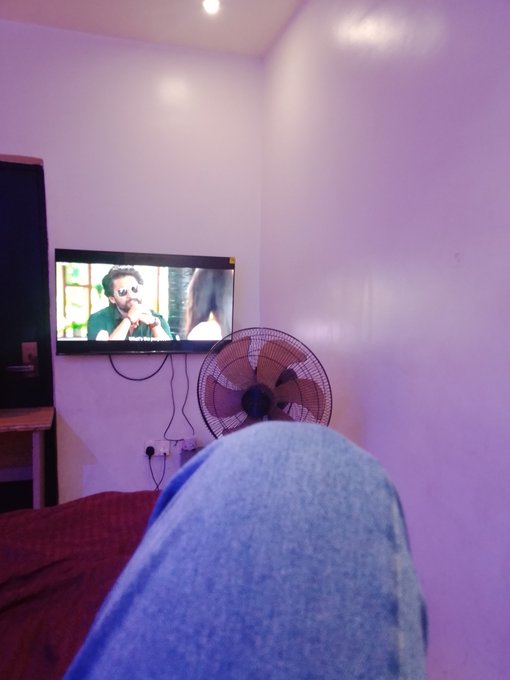 A first-person perspective photo showing the photographer's legs in blue jeans propped up in front of a wall-mounted TV playing a video, with a black standing fan nearby in a purple-lit room.