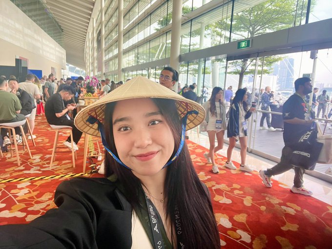 Selfie of a smiling woman wearing a traditional Vietnamese conical hat (nón lá) inside a large convention hall with other attendees.