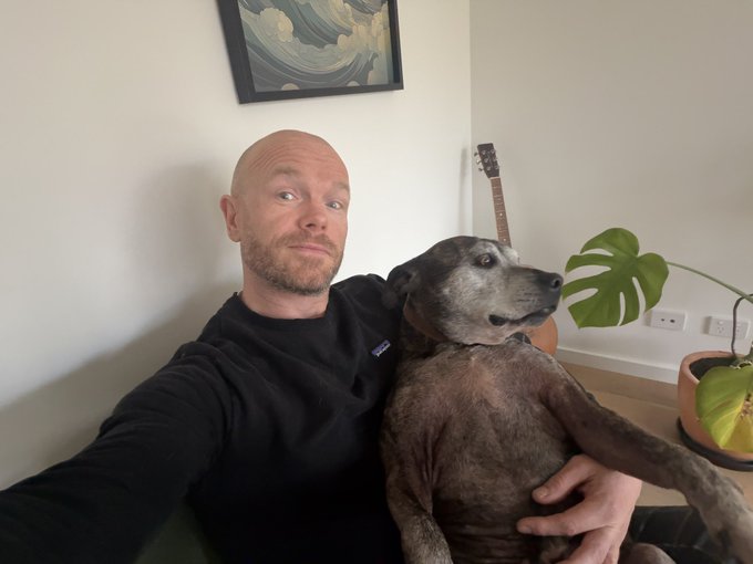 A bald man in a black sweatshirt takes a selfie while holding a gray pit bull-type dog, with a framed wave artwork, guitar, and monstera plant visible in the background.