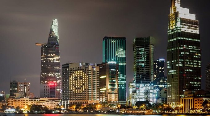 A nighttime cityscape of Ho Chi Minh City (Saigon) featuring the illuminated Bitexco Financial Tower and other modern skyscrapers along the Saigon River.