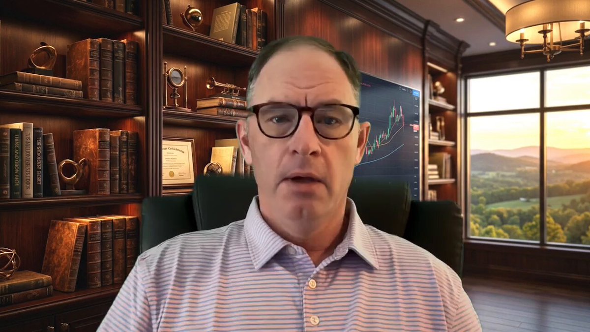 A man wearing glasses sits at a desk with a window behind him, looking directly into the camera. He has a serious expression on his face as he looks off-camera. The background features bookshelves filled with various books, suggesting that this is likely an office or study area. There are also some pictures hanging on the wall above the man's head.