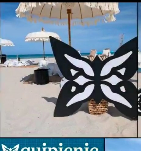 A black and white butterfly-shaped cutout stands on a white sand beach with white fringed umbrellas and blue ocean in the background.