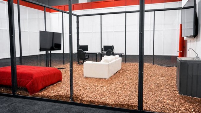 An interior display of a glass-walled room featuring a red bed, white couch, wall-mounted TV, and a desk with computers, set on a textured brown floor with a red accent ceiling.