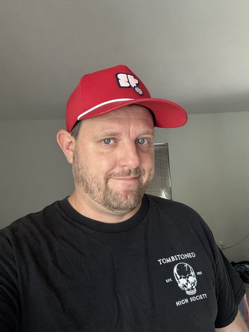 A selfie of a man wearing a red UFC cap and a black 'TOMBSTONED HIGH SOCIETY' t-shirt featuring a skull graphic, standing indoors.