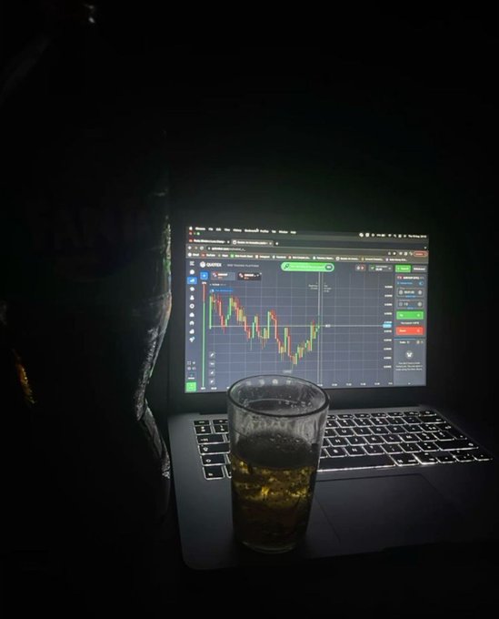 A glass of beer sits on a laptop keyboard in the dark, with a cryptocurrency trading chart displayed on the screen showing red and green candlestick patterns. The image reinforces the post's message about investing in crypto by juxtaposing leisure with the late-night reality of trading digital assets.