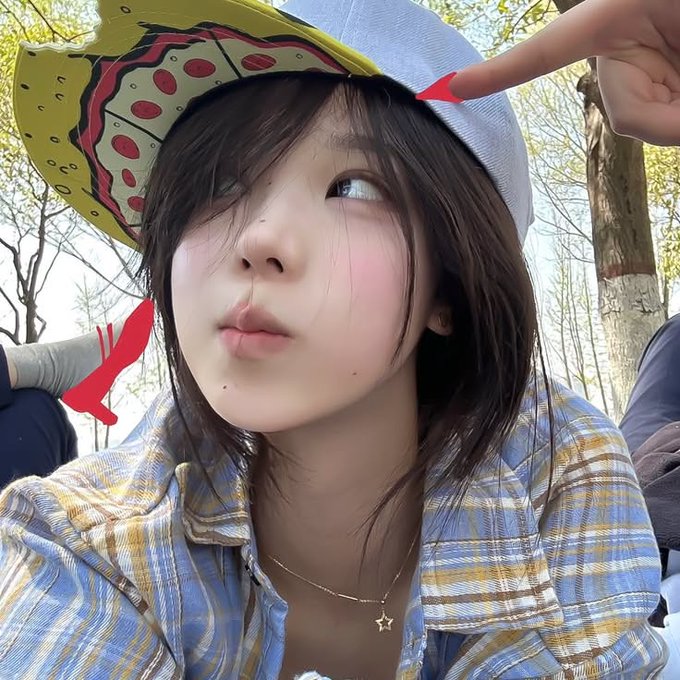 A close-up selfie of a young woman with dark hair wearing a yellow patterned cap and a blue plaid shirt, looking off to the side with her finger pointed at her temple, outdoors with trees in the background.
