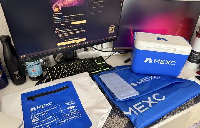 A collection of MEXC-branded merchandise, including a cooler box, pouch, drawstring bag, and what appears to be merchandise packaging featuring "USDT/BTC" and "1,000 BTC Campaign," is displayed on a desk alongside a monitor showing a MEXC Liquidity leaderboard. The image showcases the crypto exchange's promotional products as referenced in the post thanking MEXC and acknowledging contributors @BreezeYuan and @SouBolado.