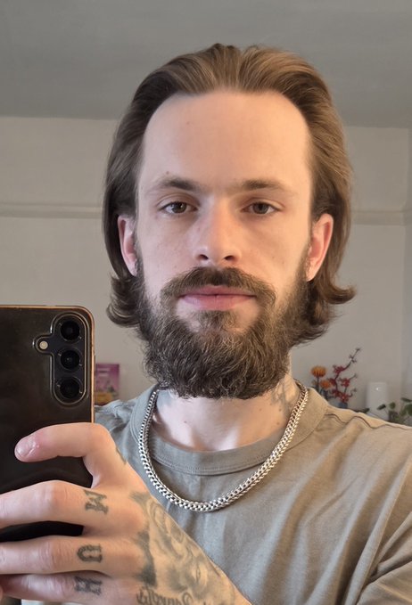 A man with shoulder-length wavy brown hair and a full beard takes a mirror selfie using a black smartphone, with visible tattoos on his hand and the letters "H" and "R" inked on his fingers. His noticeably grown-out hair directly illustrates the post's claim of a 239-day hair-growing journey tied to XRP reaching $10.