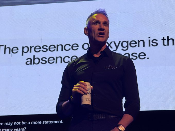 A man on stage holds a beverage can while speaking in front of a slide that partially reads 'The presence of oxygen is th... absence... ase.'