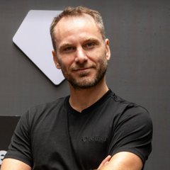 A portrait of a smiling man with short brown hair and a beard, wearing a black t-shirt with his arms crossed, standing in front of a dark wall with a geometric logo.