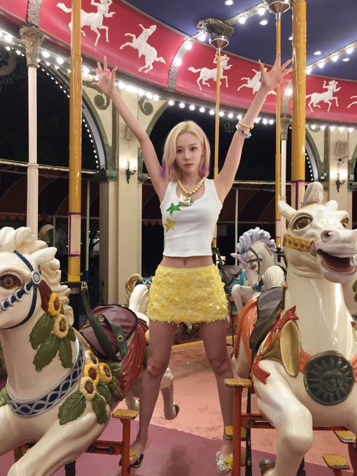 A blonde woman poses with arms raised on a decorative carousel, wearing a white crop top with stars and a yellow skirt.
