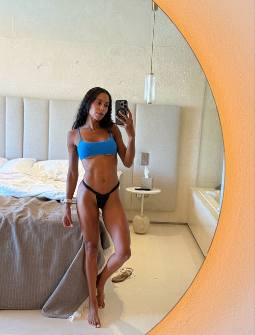 A woman in a blue bikini top and black bottoms takes a mirror selfie in a modern bedroom with a textured wall and round mirror.