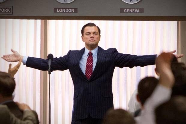 Leonardo DiCaprio as Jordan Belfort stands with arms outstretched in a pinstripe suit before an audience, with city name placards ('LONDON', 'GENEVA') visible on the wall behind him.