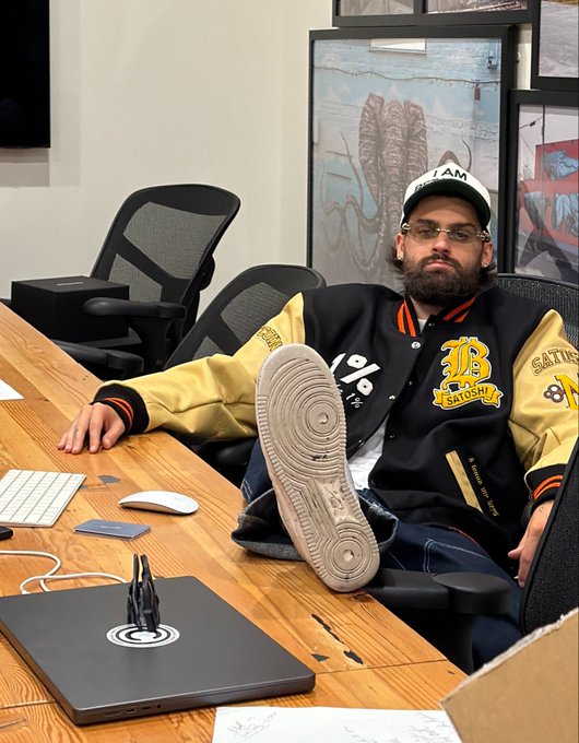 A bearded man wearing a "WILLIAM" cap and a black and yellow "B SATOSHI" varsity jacket leans back in a rolling chair at a conference table, with his sneaker propped up to show the sole. Framed "Solana" artwork in the background ties into the "SOLANA SUMMER" theme, suggesting a relaxed, confident attitude during the crypto season.