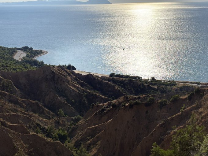 A scenic landscape showing eroded, layered clay cliffs descending toward a sparkling sea with sunlight reflecting off the water and a winding coastal road visible on the left.