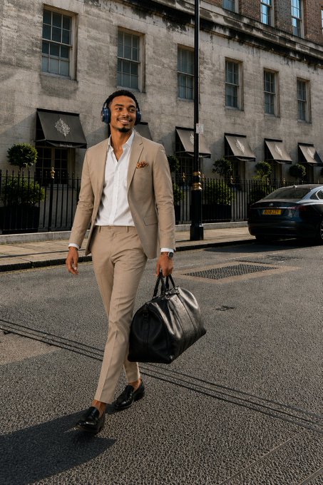 A well-dressed man in a beige suit with headphones around his neck walking down a city street while carrying a black duffle bag.