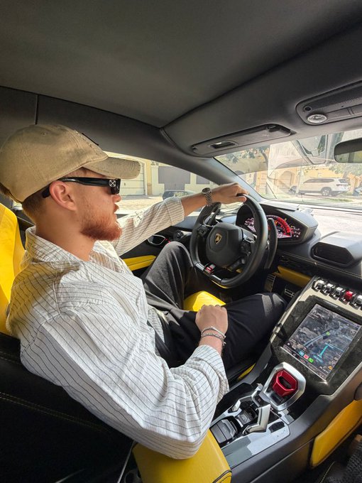 Interior shot of a young man with a reddish beard wearing a beige cap, sunglasses, and a striped shirt driving a yellow Lamborghini, with the navigation screen and steering wheel visible.