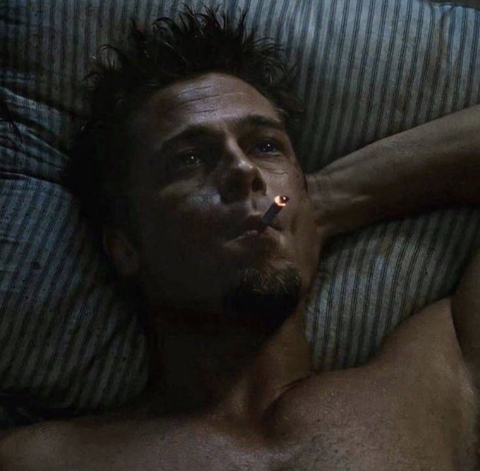 The image features a shirtless man lying down with his eyes closed, holding a lit cigarette between them. The background of the photo shows a bed with blue stripes, which contrasts with the dark hair of the man. The caption reads "Me fully exposed to the only asset on the entire planet that hasn't gone up in the last year." This phrase suggests that the image is meant to be humorous or satirical, as it implies that the man's exposure and lack of change are exaggerated for comedic effect.