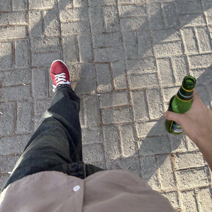 A first-person view looking down at red sneakers, jeans, and a hand holding a green glass beer bottle while walking on a paved stone surface.