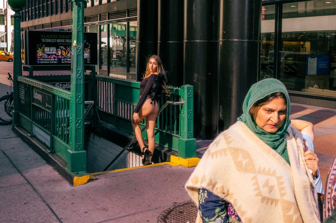 Two women, one in a revealing outfit and the other in traditional headwear, pass each other near a New York City subway entrance, perhaps symbolizing different worlds intersecting. The sign "ALAMO DRAFTHOUSE" is visible.