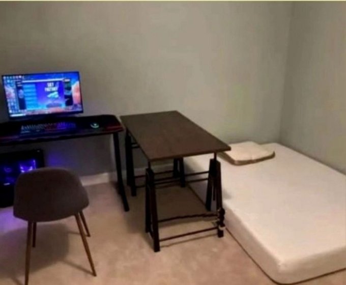 A minimalist room setup featuring a gaming desk with monitor displaying "GEF" on screen, paired with a simple folding table, a basic chair, and a bare mattress on the floor. The sparse, starting-from-scratch arrangement of essential furniture embodies the "it's okay to start like this" sentiment, showing that humble beginnings with just the basics can still create a functional living and gaming space.