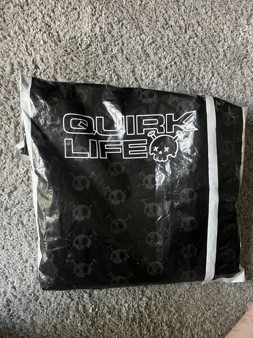 A black glossy Quirk Life themed package or bag with repeated skull logos and a bomb logo, resting on a gray carpet.