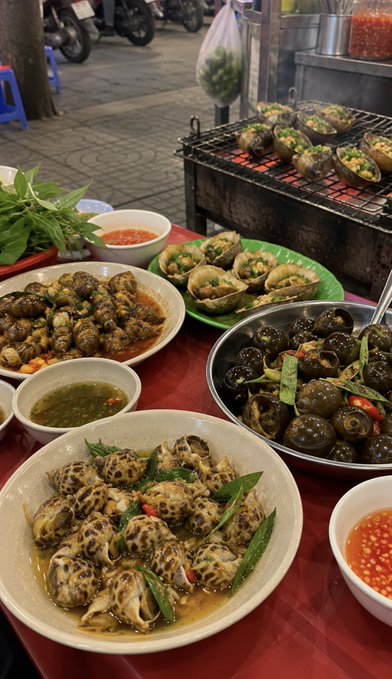 A Vietnamese street food scene displaying various plates of prepared snails and shellfish dishes with herbs and dipping sauces on a red table, with a grill in the background cooking more snails.