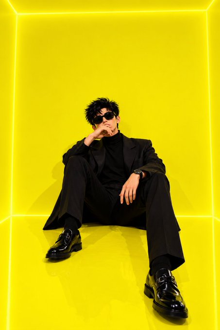 A stylish man in a black suit and sunglasses sits on the floor in a neon-yellow cube-shaped room.