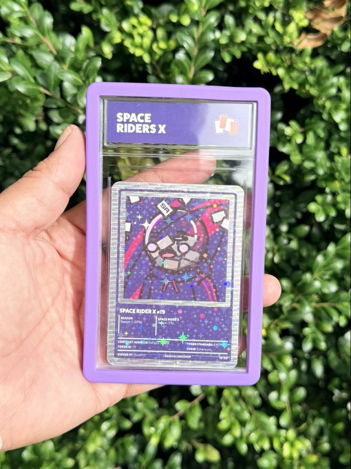 A hand holds a graded "Space Riders X #19" trading card encased in a clear slab with a vibrant purple protective guard, set against a backdrop of green foliage. The card features a colorful cosmic-themed illustration and matches the caption's reference to the Space Riders X NFT collection, with the purple guard complementing the card's celestial aesthetic.