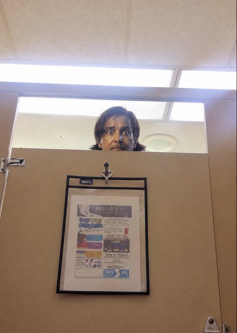 A woman peeks over the top of a bathroom stall door, looking down at a framed wall poster that displays various drug awareness flyers.