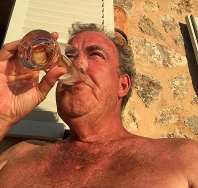 A selfie of an older shirtless man drinking from a wine glass, with sunlight casting shadows on his face against a stone wall background.