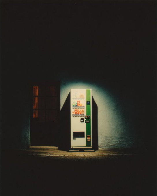 A moody, atmospheric photograph of a green and white vending machine illuminated in the dark, standing beside a dimly lit door.