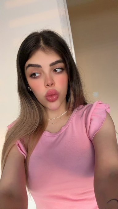 A young woman in a pink top with bow-tied shoulder straps makes a kissing face at the camera, wearing layered necklaces and small hoop earrings.