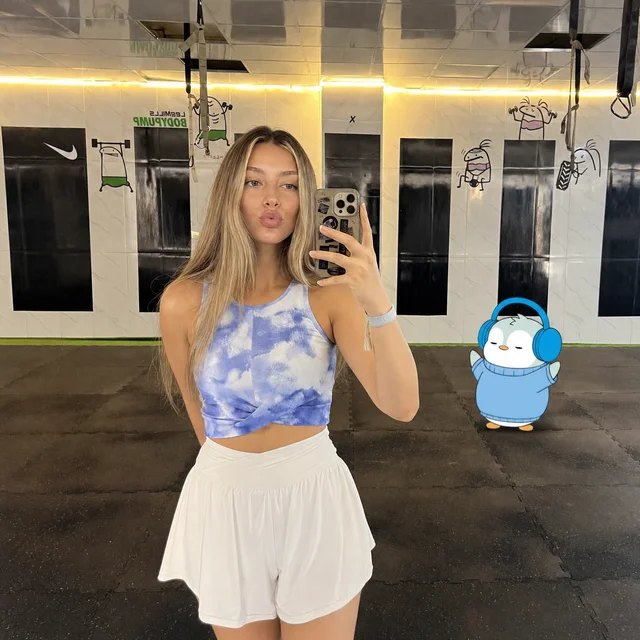A woman with long blonde hair in a blue tie-dye top and white shorts taking a mirror selfie in a gym, with a cartoon penguin sticker in the corner.