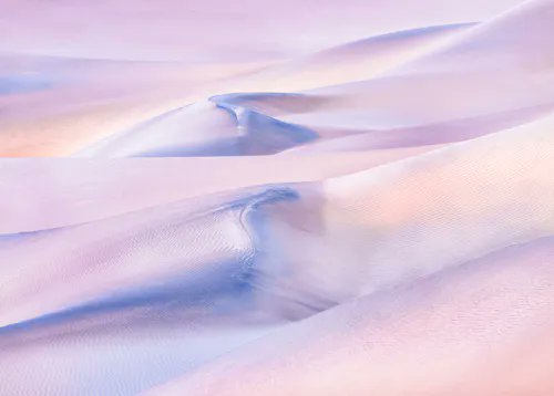 An aerial view of undulating sand dunes with a soft pink and white color palette.