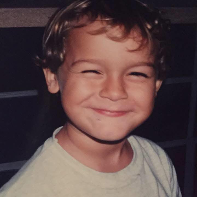 A close-up photograph of a young boy with curly blonde hair smiling warmly at the camera, wearing a white t-shirt.