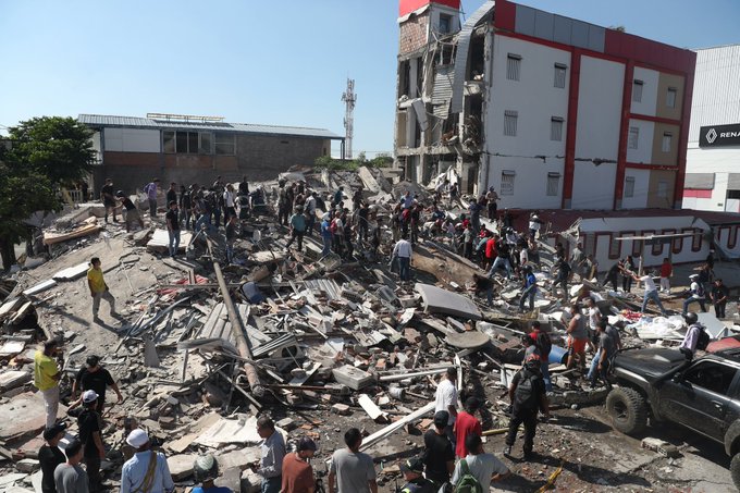 First responders and civilians sift through the massive rubble of a collapsed building following a powerful earthquake, while a damaged "RENA" sign is visible on the partially destroyed structure in the background. The image depicts the devastating aftermath in western Colombia, illustrating the urgent need for the verified crypto donation relief efforts mentioned in the post.