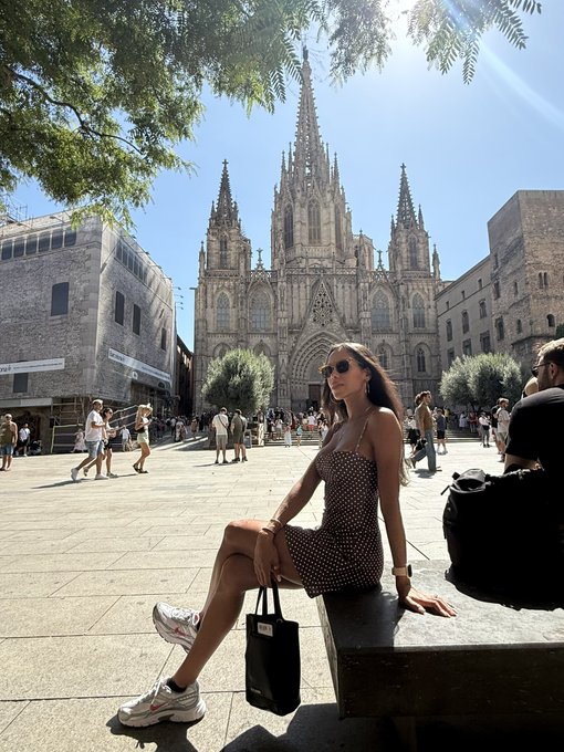 A woman in a polka-dot dress and sunglasses sits on a stone bench in a sunlit plaza, posing in front of a towering Gothic cathedral, likely Barcelona Cathedral, with other tourists and buildings visible in the background.