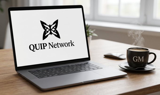 The image shows an open laptop computer sitting on top of a wooden desk, with a cup of coffee nearby. The laptop screen displays the Quip Network logo, indicating that the user might be working or studying for a project related to the Quip Network. The text in the image suggests that the Quip Network is focused on building protection for a quantum future and aims to create a network where everyone can thrive.