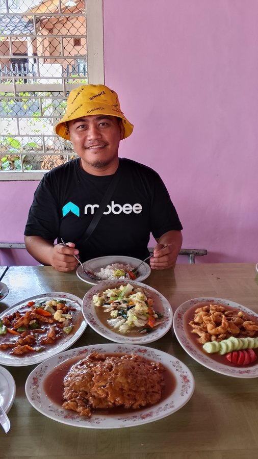 A man wearing a yellow bucket hat and a black 'mobee' T-shirt sits at a table set with various dishes of Chinese food in front of a pink wall.