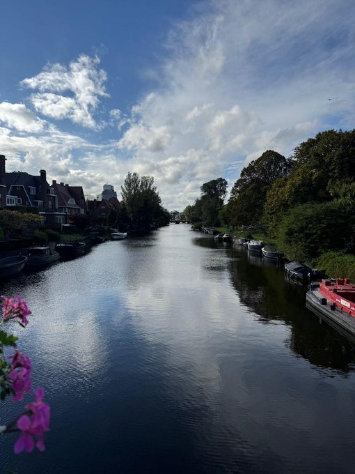 A scenic photograph of a calm canal flanked by trees and houses under a bright blue sky with scattered clouds, with small boats moored along the banks and pink flowers in the foreground.