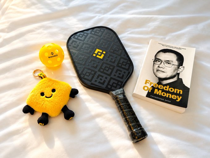 A flat-lay photo on white sheets showing a yellow Binance golf ball, a yellow smiley plush keychain, a black and gold pickleball paddle, and a book titled 'Freedom of Money' by Changpeng Zhao.
