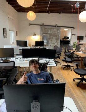 A young man sitting at a computer in a bright open-plan office with hanging spherical lights and wooden floors.