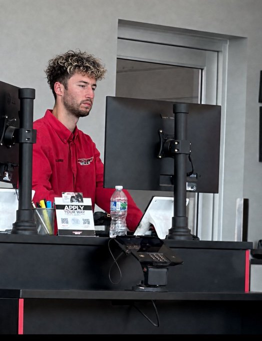 A young man with curly hair wearing a red shirt stands behind a desk with two computer monitors, a water bottle, and a phone on a payment terminal.