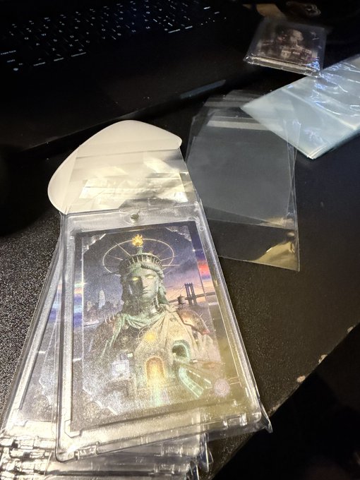 A stack of trading cards in plastic sleeves, with the top card featuring an artistic image of the Statue of Liberty and the Statue of Liberty's crown merged into a single design, sitting on a dark desk near a keyboard.